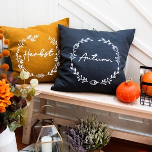 Könnte beinhalten: Zwei dekorative Kissen mit den Worten "Herbst" und "Autumn" in weißer Schrift auf einem dunkelgrauen und senfgelben Hintergrund. Die Kissen liegen auf einer hellen Holzbank mit einer Blumenvase und Kürbissen im Hintergrund.