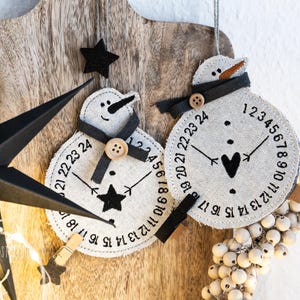 May include: Two snowman-shaped Christmas ornaments with clock faces, black scarves, and button details. The ornaments are made of a light-coloured fabric and feature black numbers and hands. A black star and a decorative star are also present.