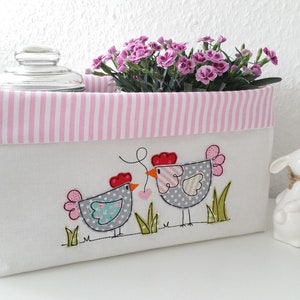 May include: A white fabric storage box with pink and white stripes around the top. The box has an embroidered design of two gray chickens with red combs and a heart between them. The box is sitting on a white surface next to a white ceramic rabbit.
