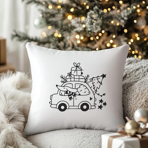 Op de afbeelding: Een wit kussen met een zwart-witte illustratie van een auto die een kerstboom en cadeaus vervoert. De auto heeft een hond op de bestuurdersstoel die een sjaal draagt.