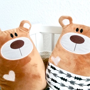 Pu&ograve; includere: Due cuscini di peluche marroni a forma di orso con volti ricamati e cuori bianchi. Un cuscino ha un motivo a frecce bianche e nere sul fondo.