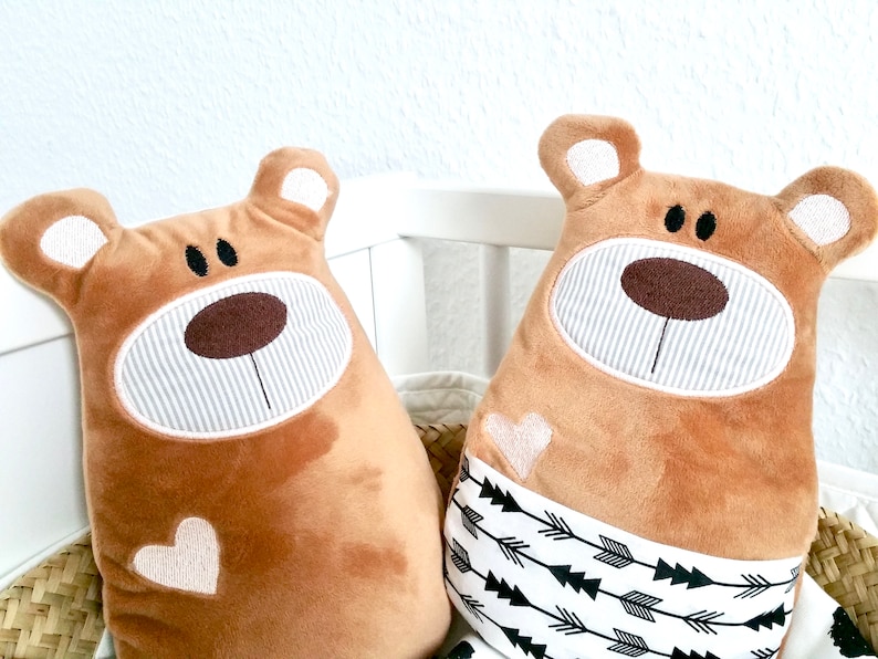 Pu&ograve; includere: Due cuscini di peluche marroni a forma di orso con volti ricamati e cuori bianchi. Un cuscino ha un motivo a frecce bianche e nere sul fondo.