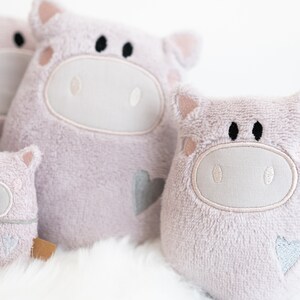 Puede incluir: Tres juguetes de peluche de cerdo rosa de diferentes tama&ntilde;os. Cada cerdo tiene una cara bordada blanca con ojos negros y un coraz&oacute;n en un lado.