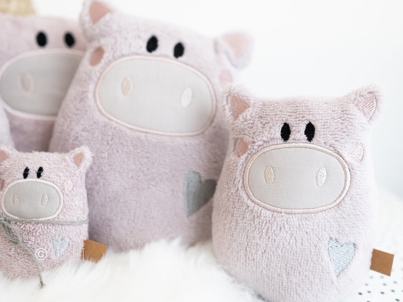 Puede incluir: Tres juguetes de peluche de cerdo rosa de diferentes tama&ntilde;os. Cada cerdo tiene una cara bordada blanca con ojos negros y un coraz&oacute;n en un lado.