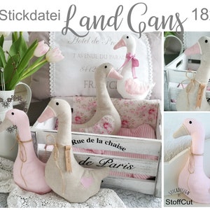 May include: Three plush goose-shaped cushions in pink and beige fabric with a heart detail. The cushions are arranged in a white wooden crate with a pink and white striped liner. The crate has a sign that reads "Rue de la chaise de Paris".