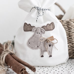 May include: A white fabric drawstring bag featuring a gray felt moose appliqué with brown antlers. The bag is tied at the top with black and white twine. A small brown paper tag is attached to the moose with a thin jute string bow.