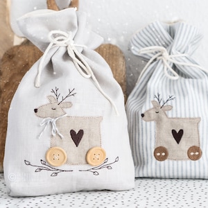 Puede incluir: Dos bolsas de regalo de lino blanco con un diseño de reno. La bolsa de la izquierda tiene un corazón marrón y la bolsa de la derecha tiene un patrón de rayas azules y blancas.