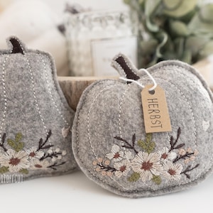 May include: Two grey felt pumpkins with embroidered flowers and a brown tag that says "HERBST" on one of them.
