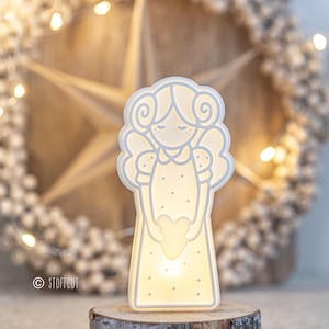 May include: A white, illuminated angel-shaped night light. The angel holds a heart and has curly hair and wings. The light sits on a wooden base, with a star and wreath in the background. The light is approximately 6 inches tall.