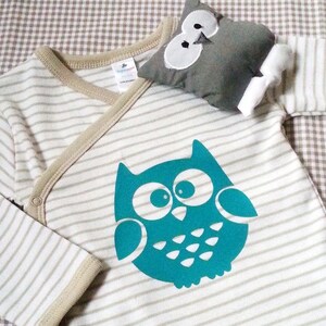 Peut inclure: Un body de b&eacute;b&eacute; &agrave; rayures beiges et blanches, orn&eacute; d'un motif de hibou turquoise. Un jouet en peluche en forme de hibou gris assorti est pos&eacute; dessus. Le body a une fermeture crois&eacute;e et une bordure beige.