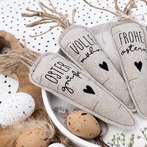 Könnte beinhalten: Vier handgefertigte Karotten-Dekorationen aus Burlap mit gestickten Ostergrüßen. Die Grüße lauten "Oster grüße", "volle  freude", "Frohe Ostern" und "Happy Easter".