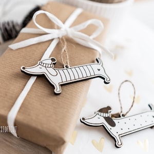 May include: Two wooden dachshund ornaments with white and black paint. One dachshund is wearing a hat and has stripes, the other has polka dots. The ornaments are hanging from a brown gift bag with a white ribbon.