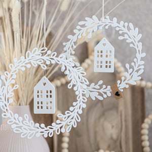 Op de afbeelding: Twee witte, bladvormige kransen met kleine, huisvormige ornamenten die erin hangen. De huizen hebben uitgesneden ramen en een hartvormige opening. Een klein label met een hart is aan één krans bevestigd. De kransen worden opgehangen aan dunne touwtjes.