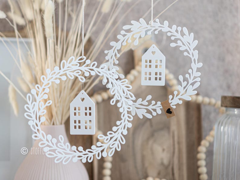 May include: Two white, leaf-shaped wreaths with small, house-shaped ornaments hanging inside. The houses have cut-out windows and a heart-shaped opening at the top. A small tag with a heart is attached to one wreath. The wreaths are suspended by thin strings.