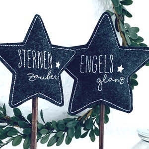 May include: Two gray felt stars on sticks with white embroidery. The left star says "Sternen Zauber" and the right star says "Engels Glanz".