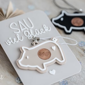 Könnte beinhalten: Eine hellgraue Karte mit dem Schriftzug "SAU viel Glück" in weißer Schrift, mit einem beigefarbenen schwein-förmigen Schlüsselanhänger mit einer Münze. Ein schwarzer Schweine-Schlüsselanhänger ist ebenfalls sichtbar. Dekorative Konfetti sind verstreut.