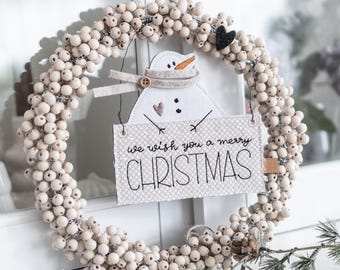 May include: A Christmas wreath made of cream-colored berries, with a snowman cutout and a sign that reads "we wish you a merry CHRISTMAS." The wreath has a small black heart and a pinecone accent.