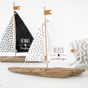 May include: Two miniature sailboats made from driftwood with white fabric sails. One sail has black polka dots and the text "Beach Cottage" with a heart. The other sail has black polka dots and the text "Heimat Hafen" with a heart.