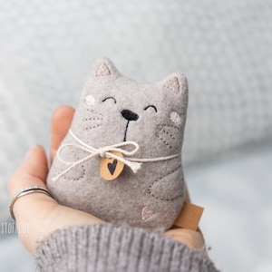Könnte beinhalten: Ein graues Filz-Katzen-Plüschtier mit gestickten Augen, einer schwarzen Nase und einem rosa Herz auf der Brust. Die Katze hält ein kleines braunes Schild mit einem Herz darauf.