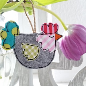May include: A gray felt chicken ornament with a pink and white striped head, a red and white striped hat, and a green and yellow polka dot tail. The ornament is hanging from a string and is in front of a vase of pink tulips.