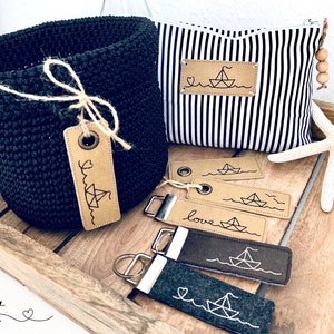 Peut inclure: Un ensemble de porte-clés sur le thème nautique et un panier en crochet noir avec une étiquette en cuir marron qui dit "love" avec un petit graphique de voilier. Les porte-clés présentent un petit graphique de voilier et le mot "love" sur une étiquette en cuir marron. Les porte-clés sont sur une surface en bois avec une étoile de mer blanche.
