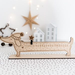 May include: Wooden dachshund-shaped advent calendar with numbers 1-24. The dog wears a hat and has a star-shaped marker. The calendar is on a wooden base, with a dotted background and decorative elements.