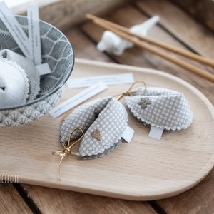 May include: Two gray and white fabric fortune cookies with gold ribbon ties. One cookie has a heart embroidered on it, and the other has a four-leaf clover embroidered on it. The cookies are on a wooden tray with white paper fortune slips.