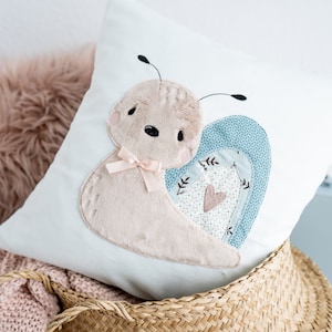 Puede incluir: Una almohada blanca con un diseño de caracol bordado en azul claro y marrón. El caracol tiene un lazo rosa y un corazón en su caparazón.