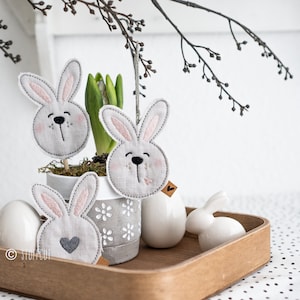Könnte beinhalten: Drei graue Filz-Hasendekorationen mit rosa Ohren und gestickten Gesichtern. Die Hasen sind in einem grauen Topf mit weißen Blumen ausgestellt. Der Topf steht auf einem Holztablett mit einem weißen Keramik-Ei und einem weißen Keramik-Hasen.