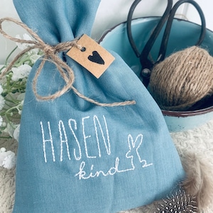 Könnte beinhalten: Eine hellblaue Stofftasche mit einem braunen Etikett, auf dem "Hasenkind" und eine weiße Hasensilhouette stehen. Die Tasche ist mit Schnur gebunden und hat ein braunes Herz auf dem Etikett.