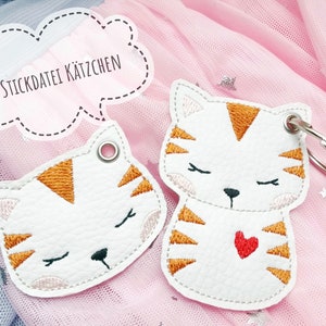 May include: Two white felt cat keychains with orange stripes and closed eyes. One keychain has a red heart on its chest. The keychains are on a pink background with white stars.