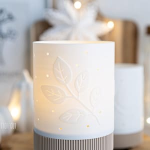 May include: A white ceramic candle holder with a leaf design and small dot cutouts. The base has a striped pattern in brown and light blue. The candle holder is lit from within, creating a warm glow. The theme is home decor.