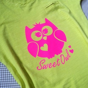 Peut inclure: T-shirt vert clair avec un motif de hibou rose et l'inscription "Sweet Owl". Le hibou a de grands yeux et un c&oelig;ur sur la poitrine. Le t-shirt a une petite poche et une bordure rose autour de l'encolure.