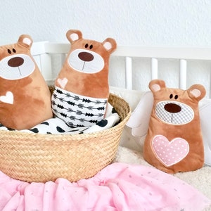 Pu&ograve; includere: Tre peluche di orso marroni con facce ricamate. Due orsi sono in un cesto intrecciato con un tessuto a motivi bianco e nero. Il terzo orso ha ali bianche e un cuore rosa con pois bianchi. Gli orsi sono su una coperta rosa e bianca.