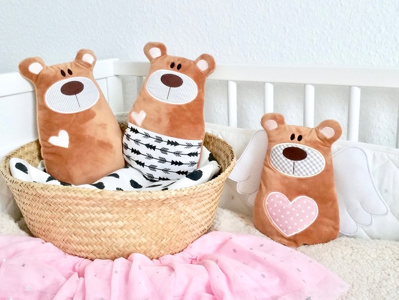 Pu&ograve; includere: Tre peluche di orso marroni con facce ricamate. Due orsi sono in un cesto intrecciato con un tessuto a motivi bianco e nero. Il terzo orso ha ali bianche e un cuore rosa con pois bianchi. Gli orsi sono su una coperta rosa e bianca.