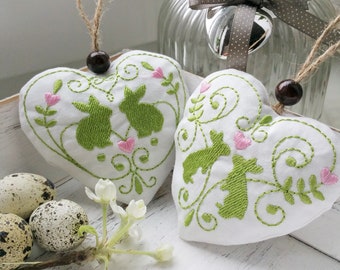 May include: Two white heart-shaped fabric ornaments with green embroidered bunnies and pink hearts. The ornaments are hanging from brown strings.