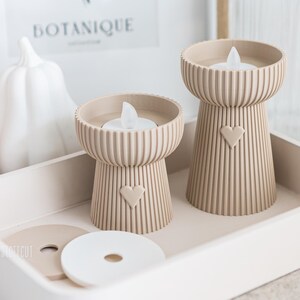 May include: Two beige candle holders with a heart design, each holding a white LED candle, sit on a white tray. The candle holders have a ribbed texture and a heart-shaped cutout. Decorative items, including a white pumpkin, are in the background.