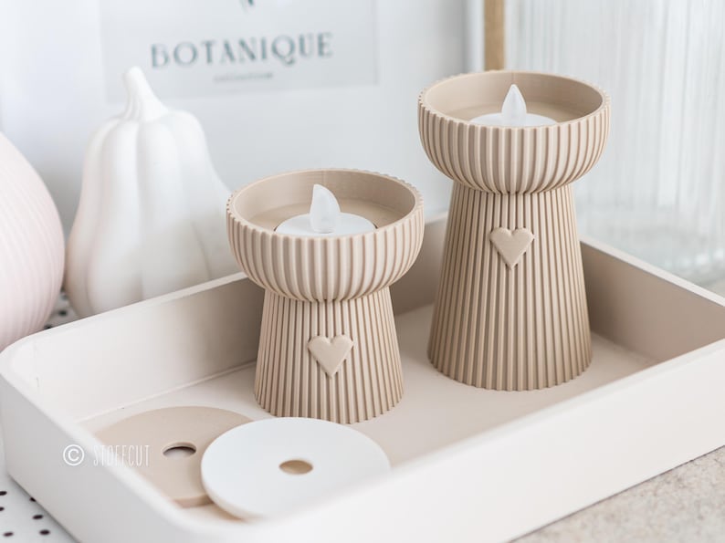 May include: Two beige candle holders with a heart design, each holding a white LED candle, sit on a white tray. The candle holders have a ribbed texture and a heart-shaped cutout. Decorative items, including a white pumpkin, are in the background.