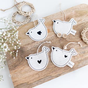 Könnte beinhalten: Vier weiße Filz-Vogel-Ornamente mit schwarzen Stickdetails. Jeder Vogel hat ein anderes Design, darunter ein Herz, ein Kreis und ein kleiner Vogel. Die Ornamente hängen an einer Holzoberfläche mit dem Text "Hand" auf zwei von ihnen.