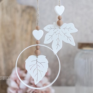 Può includere: Ornamenti bianchi a forma di foglia e cuore appesi a uno spago. Gli ornamenti sono decorati con perline di legno marrone. Una foglia è all'interno di una cornice circolare bianca. Lo sfondo è sfocato, con un vaso e piante secche visibili.