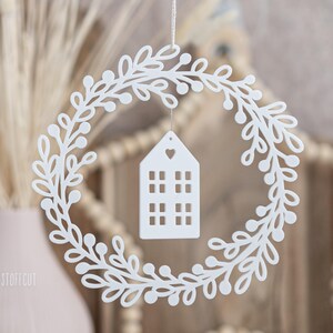 May include: A white, laser-cut wreath with a leafy design and small berries. A white house-shaped ornament with window cutouts and a heart detail hangs in the center. The wreath is suspended by a thin string, suggesting a decorative item.