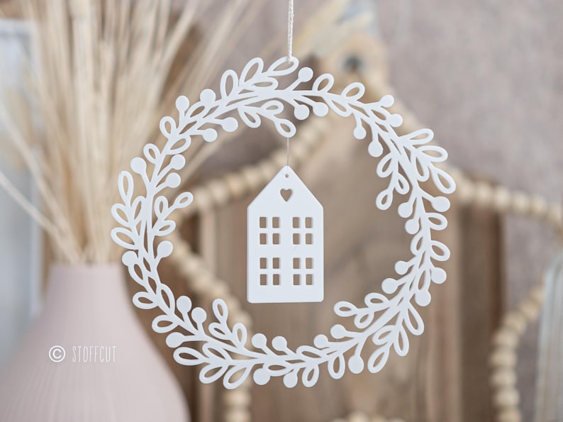 May include: A white, laser-cut wreath with a leafy design and small berries. A white house-shaped ornament with window cutouts and a heart detail hangs in the center. The wreath is suspended by a thin string, suggesting a decorative item.