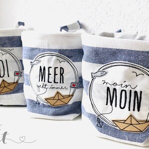 Könnte beinhalten: Drei weiße Stoffaufbewahrungskörbe mit blauen Streifen. Jeder Korb hat ein anderes gesticktes Design mit einem Papierboot und Text. Der erste Korb sagt "Ahoi", der zweite Korb sagt "Meer geht immer" und der dritte Korb sagt "moin MOIN".