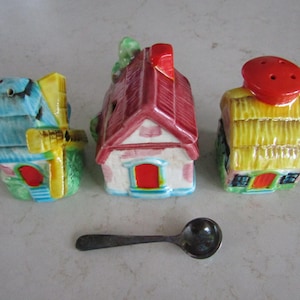 Peut inclure: Trois salières et poivrières en céramique en forme de maison avec une petite cuillère. Les salières et poivrières sont peintes de couleurs vives, dont le bleu, le rouge et le jaune. La cuillère est argentée.