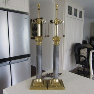 May include: A pair of gold and black table lamps with fluted columns and ornate bases. The lamps have two light bulbs each and are wired with pull chains.