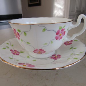 May include: A white teacup and saucer set with a delicate floral pattern. The cup and saucer are trimmed with a gold band. The floral pattern features pink flowers and green leaves.