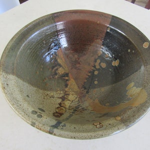 May include: A large, round ceramic bowl with a mottled glaze in shades of brown, green, and black. The bowl has a speckled, textured surface and a slightly raised rim.