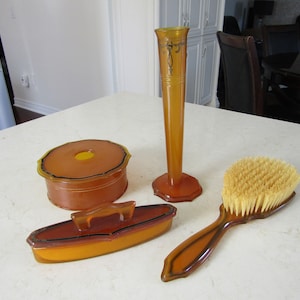 May include: A vintage amber-colored vanity set with a round powder box, a rectangular brush holder, a tall vase, and a brush with natural bristles.