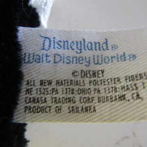 May include: Close-up of a tag with the text "Disneyland Walt Disney World" in blue. The tag also includes the Disney logo and text about materials and origin. The tag is attached to a black and white plush toy.