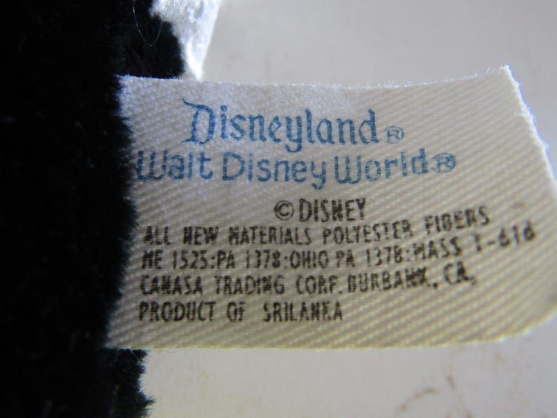 May include: Close-up of a tag with the text "Disneyland Walt Disney World" in blue. The tag also includes the Disney logo and text about materials and origin. The tag is attached to a black and white plush toy.